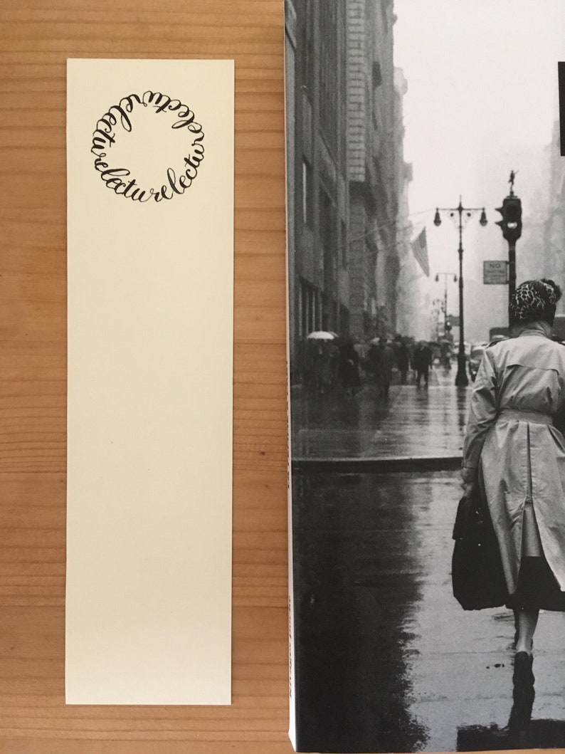 May include: A cream-colored rectangular bookmark featuring a circular design made of the repeated word "lecture" in black script, positioned next to a black and white photograph of a person in a trench coat walking down a rainy city street.