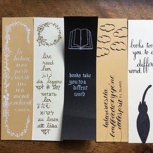 May include: Five rectangular paper bookmarks featuring various designs and text. One is tan with white floral borders and French text, one is white with multi-language text, one is black with a book icon, one is tan with clouds, and one is white with a cat.