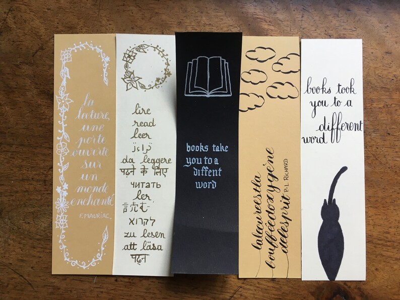 May include: Five rectangular paper bookmarks featuring various designs and text. One is tan with white floral borders and French text, one is white with multi-language text, one is black with a book icon, one is tan with clouds, and one is white with a cat.