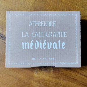May include: A brown cardboard box with white text that reads "Apprendre La Calligraphie m&eacute;di&eacute;vale De 7 A 107 Ans".