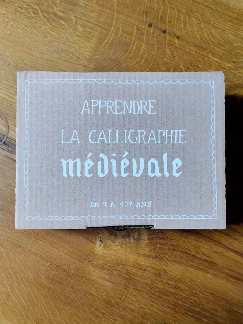May include: A brown cardboard box with white text that reads "Apprendre La Calligraphie m&eacute;di&eacute;vale De 7 A 107 Ans".