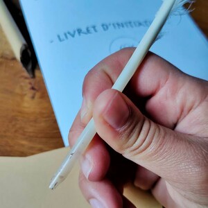 May include: A white quill pen with a feather on the end, held in a hand. The pen is resting on a white piece of paper with the text "La collection m&eacute;di&eacute;vale" and "Livret d'initiation" written on it.