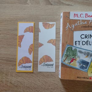 May include: Two rectangular paper bookmarks featuring a repeating pattern of illustrated golden-brown croissants. One bookmark has a yellow border, while the other is plain white. Both display the text "le Croissant since 1920" at the bottom.