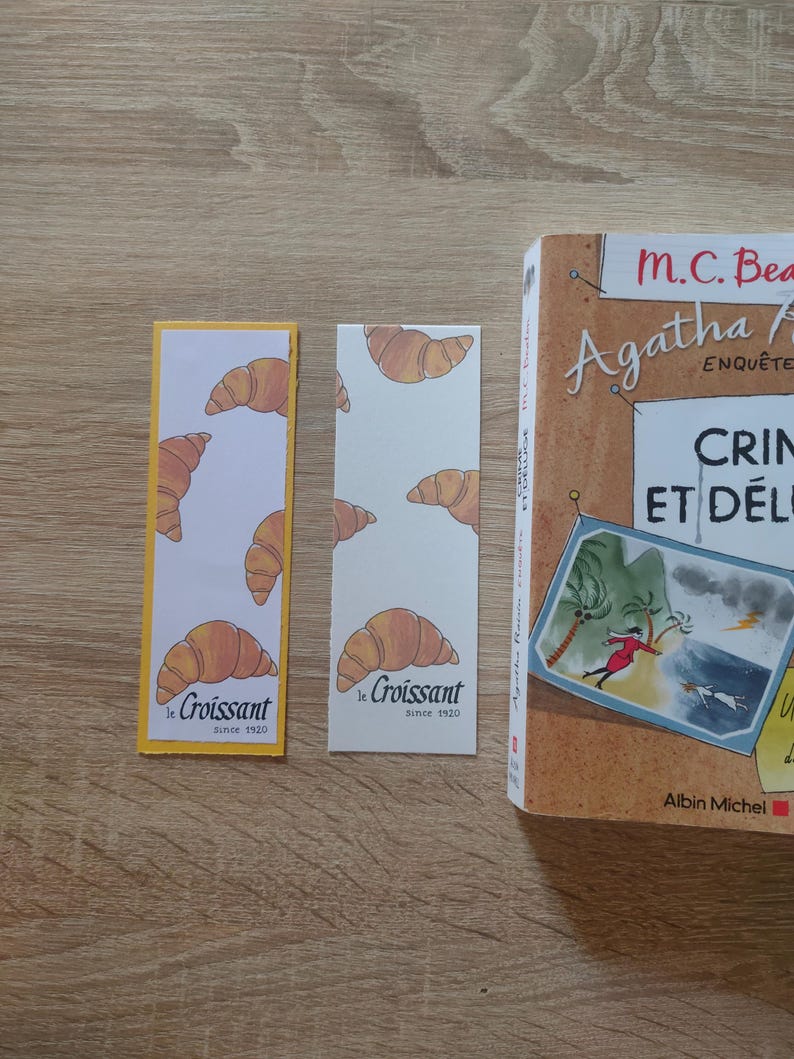 May include: Two rectangular paper bookmarks featuring a repeating pattern of illustrated golden-brown croissants. One bookmark has a yellow border, while the other is plain white. Both display the text "le Croissant since 1920" at the bottom.