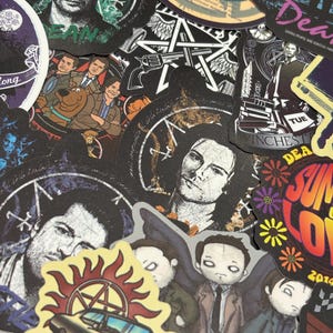 May include: A collection of Supernatural themed stickers featuring characters, symbols, and quotes from the TV show. The stickers are printed on a black background with white, red, and yellow accents. The stickers include images of the Winchester brothers, the Impala, and the show's logo.