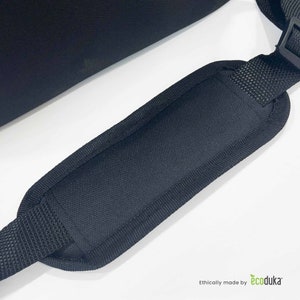 May include: Close-up of a black shoulder strap with a padded section for comfort. The strap is made of a durable, textured fabric and features a black plastic buckle. The image is set against a white background.