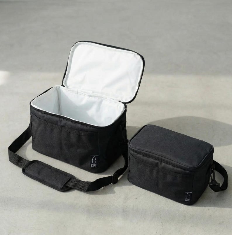 May include: Two black insulated lunch bags. One bag is open, revealing a white interior, and the other is closed. Both bags have a black shoulder strap and a small white logo on the front.