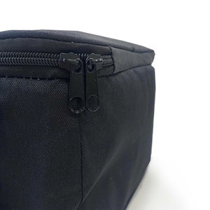 May include: Close-up of a black rectangular bag with a black zipper. The bag is made of a textured fabric and has a double zipper closure. The background is white.