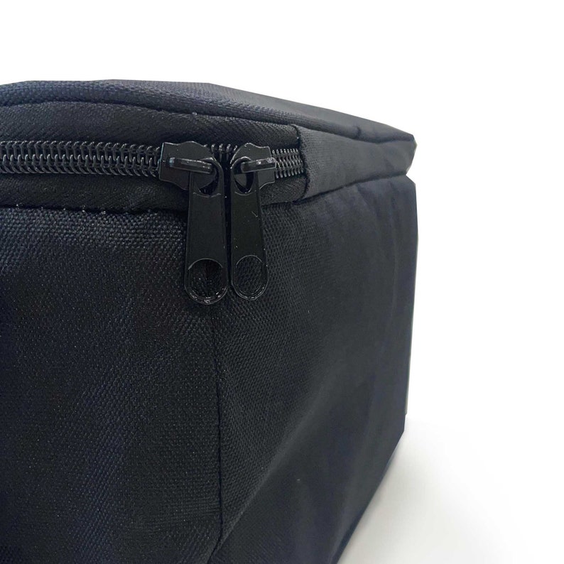 May include: Close-up of a black rectangular bag with a black zipper. The bag is made of a textured fabric and has a double zipper closure. The background is white.