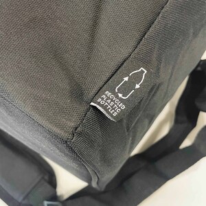May include: Black backpack with a tag that reads "RECYCLED PLASTIC BOTTLES" and a recycling symbol. The bag is made of a textured material and has a shoulder strap visible in the image.