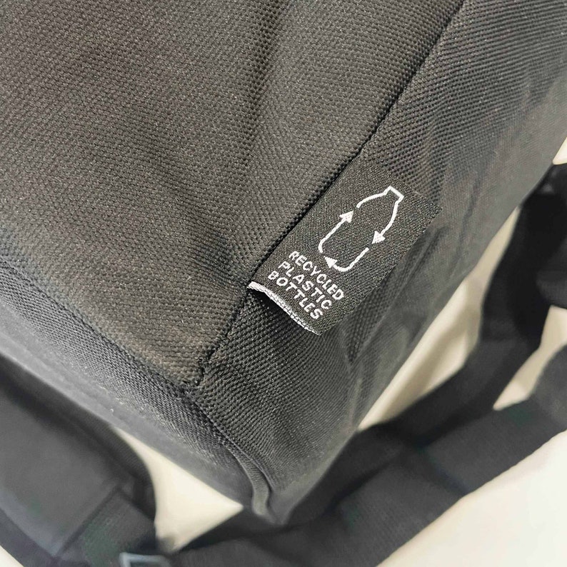 May include: Black backpack with a tag that reads "RECYCLED PLASTIC BOTTLES" and a recycling symbol. The bag is made of a textured material and has a shoulder strap visible in the image.