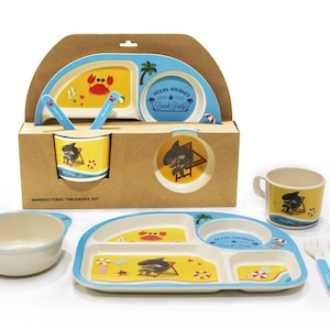 May include: A set of bamboo fibre tableware for children featuring a beach theme. The set includes a divided plate, a bowl, two cups, a spoon and a fork. The plates and cups feature a shark and crab design with the text "Ocean Journey Beach Party".