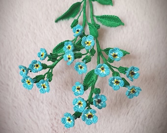 May include: A crocheted bouquet of blue forget-me-not flowers with yellow centers and green stems. The flowers are arranged in a cluster, with some flowers facing upwards and others facing downwards. The bouquet is handmade by Niran.