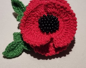 May include: A red crocheted poppy flower with a black beaded center and green crocheted leaves.