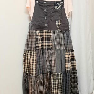 May include: A light peach-colored t-shirt layered under a dark gray denim overall dress. The dress features a bib with metal buttons, a chest pocket with a floral patch, and a tiered skirt constructed from various black, gray, and tan plaid fabrics.