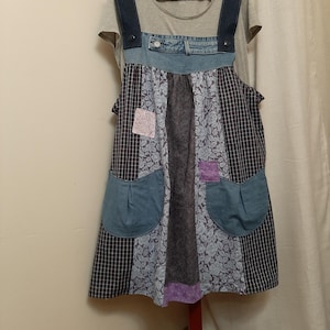 May include: A sleeveless apron dress featuring a patchwork design of blue denim, dark plaid fabric, and light blue floral print. The garment includes two large rounded pockets, a denim waistband with a button, and is layered over a gray t-shirt.