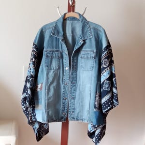 May include: A light blue denim jacket with dark blue patterned sleeves and patches. The jacket has two chest pockets and a button-up front. The sleeves feature a paisley-like design in shades of blue and white.