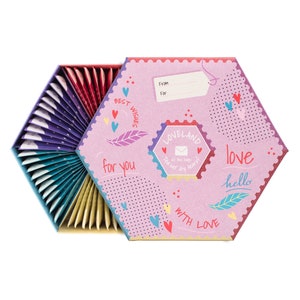 K&ouml;nnte beinhalten: Eine sechseckige Box mit einem rosa-wei&szlig;en Design mit Herzen, einer Feder und den Worten "Beste W&uuml;nsche", "f&uuml;r dich", "Loveland", "Liebe", "Hallo" und "Mit Liebe". Die Box ist ge&ouml;ffnet und zeigt einen F&auml;cher mit bunten Teebeuteln.