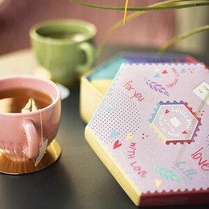 K&ouml;nnte beinhalten: Eine rosa Teetasse mit einem Teebeutel steht auf einem goldenen Untersetzer auf einem grauen Tisch. Eine violette Schachtel mit sechseckiger Form und bunten Designs ist auf dem Tisch ge&ouml;ffnet. Die Schachtel tr&auml;gt die Worte "f&uuml;r dich" und "mit Liebe". Die Schachtel ist mit Herzen, Federn und dem Wort "Liebe" verziert.