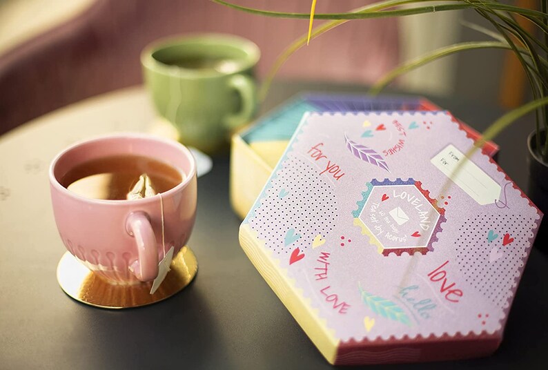 K&ouml;nnte beinhalten: Eine rosa Teetasse mit einem Teebeutel steht auf einem goldenen Untersetzer auf einem grauen Tisch. Eine violette Schachtel mit sechseckiger Form und bunten Designs ist auf dem Tisch ge&ouml;ffnet. Die Schachtel tr&auml;gt die Worte "f&uuml;r dich" und "mit Liebe". Die Schachtel ist mit Herzen, Federn und dem Wort "Liebe" verziert.