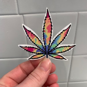 K&ouml;nnte beinhalten: Ein bunter Batik-Cannabisblatt-Aufkleber mit schwarzem Umriss.