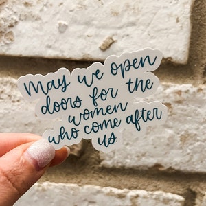 May include: A white sticker with the text "May we open doors for the women who come after us." in blue cursive lettering.