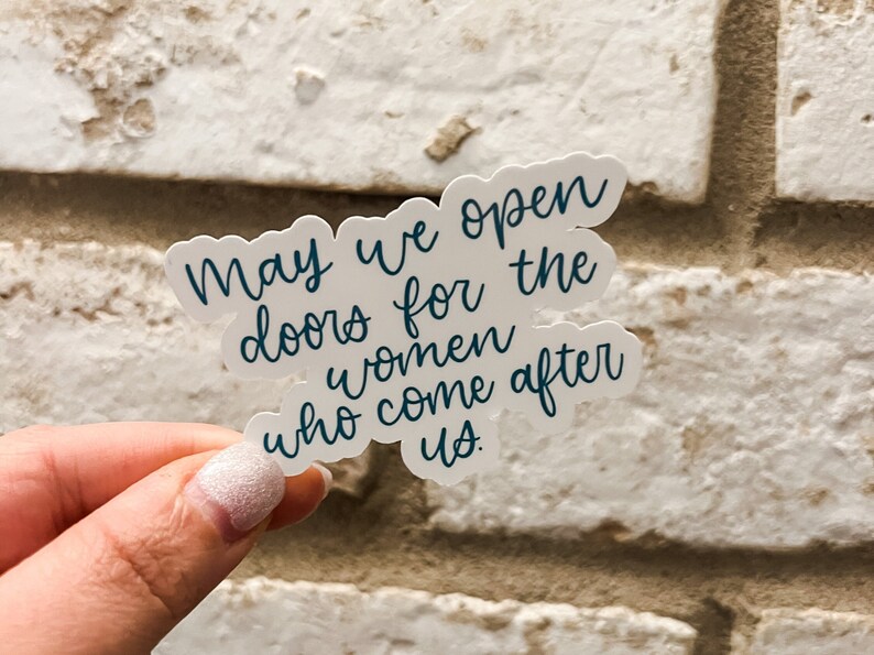 May include: A white sticker with the text "May we open doors for the women who come after us." in blue cursive lettering.