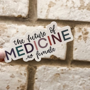 May include: A white sticker with the text "the future of MEDICINE is female" in colorful letters. The sticker is held in a hand against a white brick background.