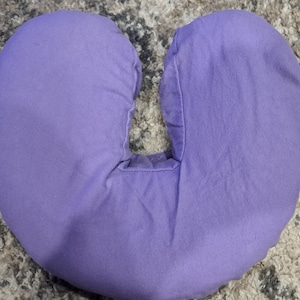 May include: A purple neck pillow with a hole in the center for the neck. The pillow is made of soft fabric and is designed to provide support and comfort.