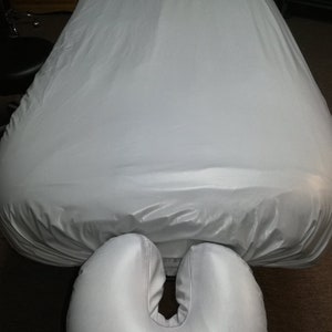 May include: A white massage table cover and face cradle. The cover is draped over the table, and the face cradle is positioned at the head of the table. The items are made of a smooth, shiny material.