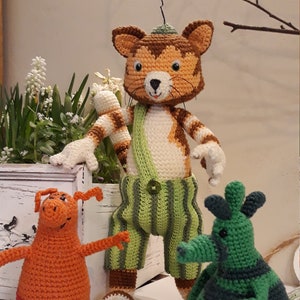 Puede incluir: Tres animales de peluche de crochet: un gato marrón y blanco con un mono verde, un cerdo naranja y una criatura rayada verde y negra.