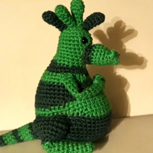 Puede incluir: Juguete de canguro de crochet verde y negro. El canguro está sentado con los brazos cruzados y la cola enroscada detrás de él.