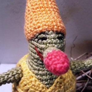 Puede incluir: Un gnomo de crochet con cuerpo verde, camisa amarilla, gorro naranja y nariz rosa.