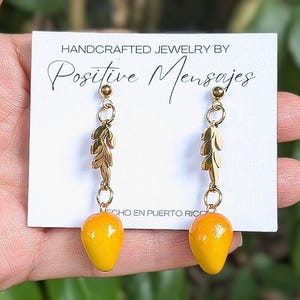 May include: A pair of handcrafted gold-colored earrings with a leaf design and a yellow mango-shaped bead. The earrings are suspended from a small gold ball and are displayed on a white card with the text "HANDCRAFTED JEWELRY BY Positive Mensajes".