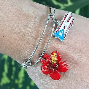 May include: A silver-toned bangle bracelet with two charms. One charm is a red hibiscus flower with yellow accents, and the other is a Puerto Rican flag. The bracelet is worn on a wrist.