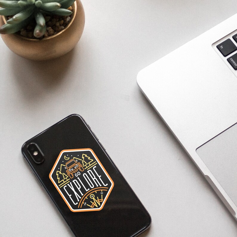 May include: A black smartphone with a sticker on the back. The sticker is a yellow and orange design with a jeep driving through a forest with the words "Go Explore" around it.