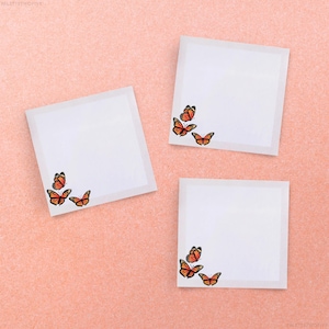 May include: Three white square sticky notes with orange butterfly designs in the corner. The sticky notes are on a light pink background.