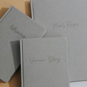 May include: Three gray linen-covered journals with different titles: "Adriana", "Mum's Recipes", and "Vance's Story".