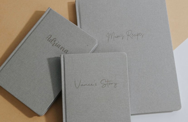 May include: Three gray linen-covered journals with different titles: "Adriana", "Mum's Recipes", and "Vance's Story".