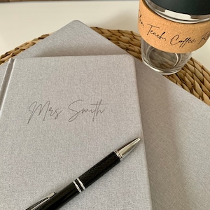 May include: Two gray journals with a pen on top of the top journal. The top journal has the name "Mrs. Smith" written on the front.