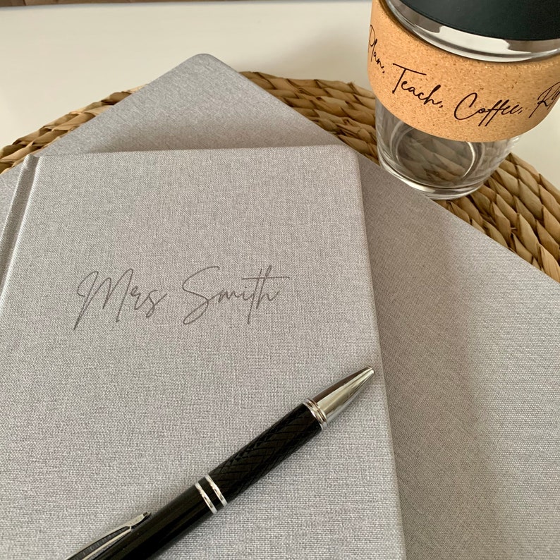 May include: Two gray journals with a pen on top of the top journal. The top journal has the name "Mrs. Smith" written on the front.