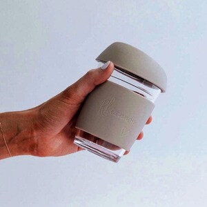 May include: A clear glass reusable coffee cup with a taupe silicone sleeve and lid. The sleeve is embossed with the name "Rhiannon" and a heart symbol. The cup is held in a hand against a white background.