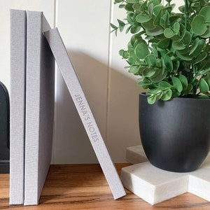 May include: Three gray linen-covered journals stacked on a wooden surface. The top journal has the text "Jenna's Notes" embossed on the cover.