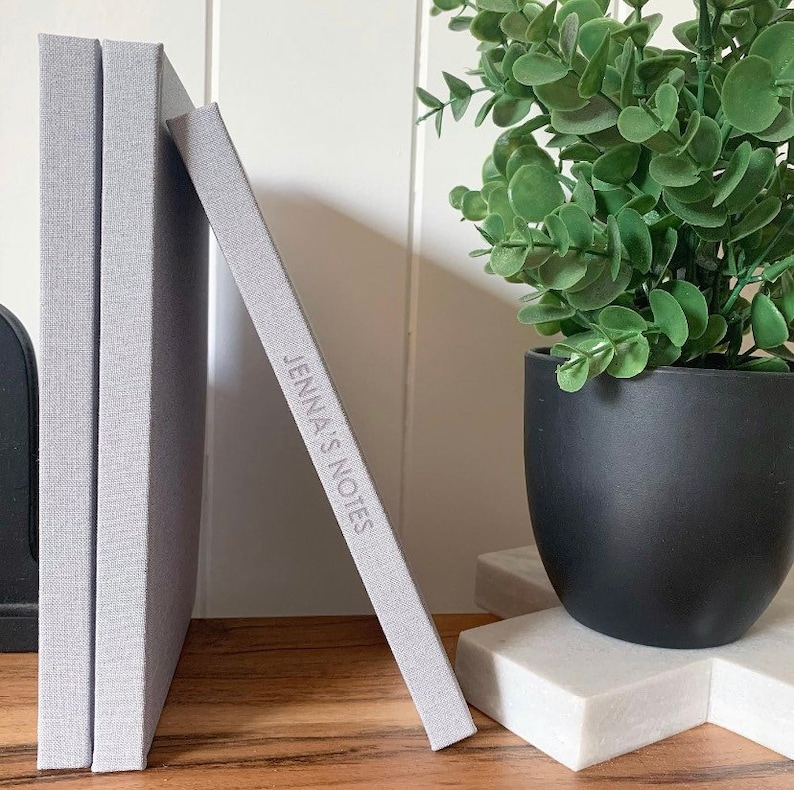 May include: Three gray linen-covered journals stacked on a wooden surface. The top journal has the text "Jenna's Notes" embossed on the cover.