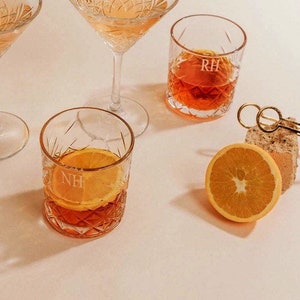 Op de afbeelding: Twee heldere glazen met gegraveerde initialen "RH" en "NH", gevuld met een donkeroranje vloeistof. Een halve sinaasappelschijf ligt op een wit oppervlak.