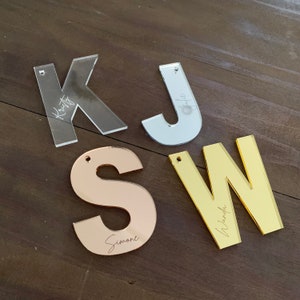 Op de afbeelding: Vier acryl letters: een doorzichtige "K" met "Kristy", een zilveren "J" met "Julie", een ros&eacute;gouden "S" met "Simone" en een gouden "W" met "Wanda". Elke letter heeft een klein gaatje om op te hangen.