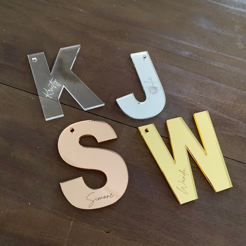 Op de afbeelding: Vier acryl letters: een doorzichtige "K" met "Kristy", een zilveren "J" met "Julie", een ros&eacute;gouden "S" met "Simone" en een gouden "W" met "Wanda". Elke letter heeft een klein gaatje om op te hangen.