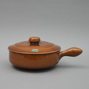 May include: A brown ceramic casserole dish with a lid and a single handle. The dish has a green label that reads "Carl Schmidt Sohn".