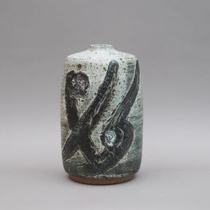 Puede incluir: Un jarrón de cerámica con un esmalte moteado blanquecino y una base marrón oscuro. El jarrón presenta un diseño abstracto y audaz en negro, creando un contraste llamativo. El jarrón es cilíndrico con una parte superior ligeramente redondeada.
