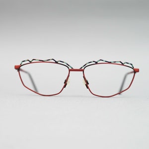 Puede incluir: Gafas de metal rojo con montura geométrica y un detalle azul y verde en la parte superior de la montura.